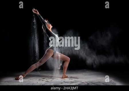 Ballerina flessibile ballerina donna ballare e cospargere la farina su sfondo nero, attraente sottile donna sottile in bodysuit nero in slow motion, gra in movimento Foto Stock
