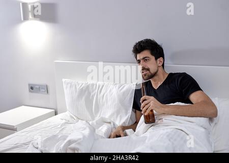 Scatto interno di giovane maschio grumpy rilassarsi in camera da letto, guardare la serie televisiva, bere alcol a letto, commentando ciò che accade sullo schermo. Bearde Foto Stock