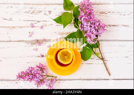 una tazza di ceramica gialla con tè e una fetta di limone su un tavolo di legno bianco con rami di belle lillacs. il concetto di una vacanza di campagna Foto Stock