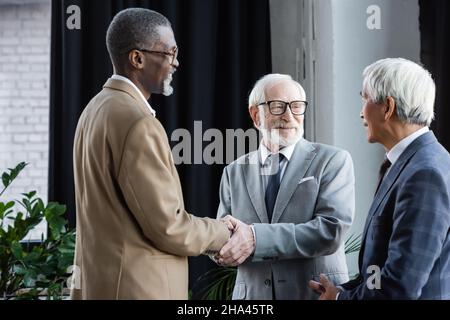 felice partner di affari interrazziali scuotendo le mani vicino ad un collega asiatico anziano Foto Stock