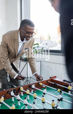 uomo d'affari afroamericano senior che gioca a calcio da tavolo con un collega sfocato Foto Stock