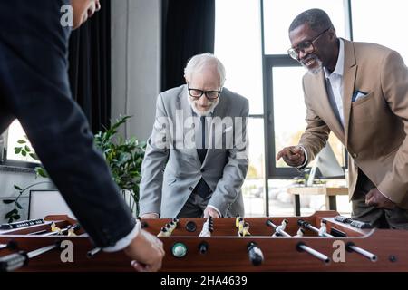 sorridente uomo d'affari afroamericano che punta con un dito mentre colleghi senior giocano a calcio balilla Foto Stock