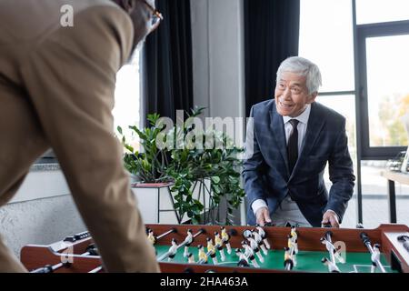 offuscato uomo d'affari afroamericano che gioca a calcio da tavolo con un collega asiatico senior Foto Stock