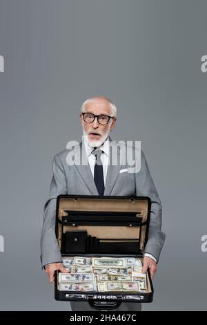 stupito uomo d'affari senior mostrando briefcase pieno di dollari isolato su grigio Foto Stock