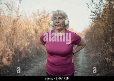 Donna anziana che corre con cuffie e smart watch in un parco in una bella natura autunnale. Natura e sport concetto di stile di vita sano. Foto Stock