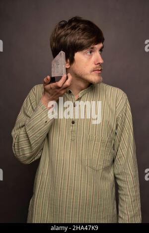 Giovane uomo bianco sottile, alto e bello, con capelli marroni che ascoltano un pezzo di cristallo in camicia a righe su sfondo grigio Foto Stock