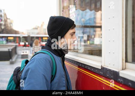 Il giovane uomo bianco bello con capelli marroni in giacca blu con cappello nero d'inverno guarda attraverso la finestra sulla strada Foto Stock