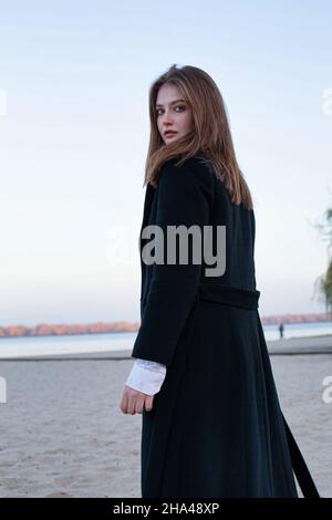 Camminando lungo dritto capelli bianchi europei donna in cappotto scuro in autunno sulla spiaggia Foto Stock