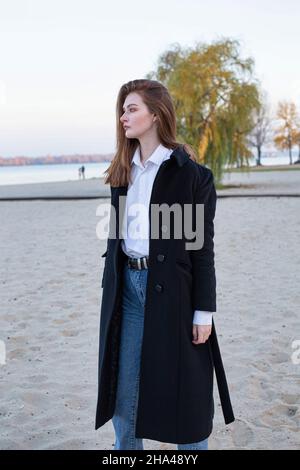 Donna europea bianca a capelli lisci lunghi in una camicia bianca in cappotto scuro in autunno sulla spiaggia Foto Stock