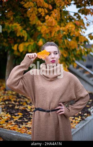 Ritratto della giovane donna bianca europea con capelli biondi in autunno tenendo la foglia gialla davanti al suo occhio Foto Stock