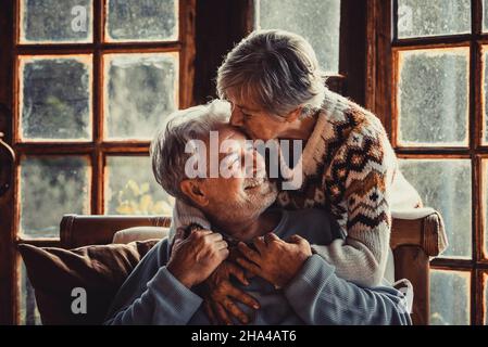 le persone anziane a casa in amore baciarsi e prendersi cura l'un l'altro. felice rapporto uomo e donna maturi insieme. vecchio maschio seduto sul divano e donna anziana abbracciandolo con cura Foto Stock
