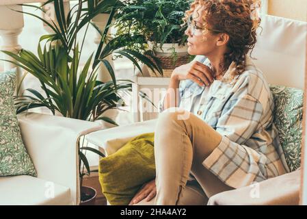 donna adulta rilassata e attraente che strega sulla sedia a casa fuori sul balcone guardando via e pensare. bella donna rilassarsi e riposare siediti. hipster ritratto casual stile Foto Stock