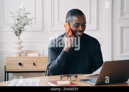 Joyful African maschio sviluppatore siede alla scrivania guarda lo schermo del computer mentre parla su smartphone Foto Stock
