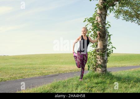 Tutta la lunghezza di sportivo adulto sicuro con capelli biondi lunghi in activewear riscaldamento e guardando lontano vicino albero prima di correre su percorso stretto in i Foto Stock