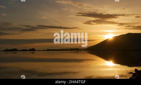 grecia,isole greche,isole ionie,lefkada o lefkas,lefkada città,capitale,luce del mattino,alba,alba,sole dietro la collina,raggi di sole,cielo azzurro e arancione con le nuvole grigie,si riflettono nell'acqua Foto Stock