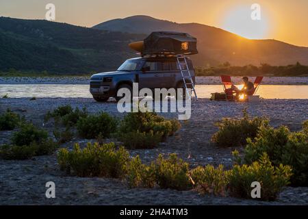 Albania, Europa meridionale, il giovane uomo si siede di fronte a un veicolo fuoristrada con tenda sul tetto e gode il tramonto Foto Stock