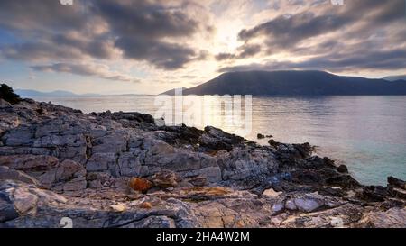 grecia,isole greche,isole ionie,cefalonia,fiskardo,umore del mattino,cielo parzialmente nuvoloso,immagine hdr,vista dell'alba dietro le nuvole grigie,rocce colorate in primo piano,raggio del sole sul mare,ithaca sotto le nuvole Foto Stock