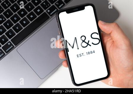 Marks e Spencer app sullo schermo di smartphone in mano mans con un laptop sullo sfondo. Acquisti online tramite telefono cellulare. Novembre 2021, San Francisco, USA Foto Stock