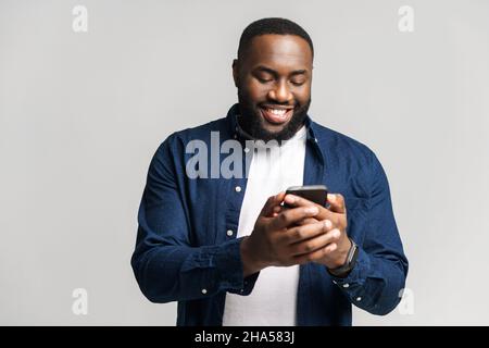 Uomo ottimista afro-americano che indossa una maglietta casual testing sullo smartphone, texting, ragazzo nero sorridente godendosi utilizzando la nuova app mobile isolata in grigio Foto Stock