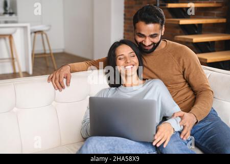 Una coppia molto gioiosa che ama trascorrere il tempo libero online con un notebook a casa. Uomo e donna indiana che guardano lo schermo del computer portatile mentre si siede sul divano e si guardano film comici Foto Stock