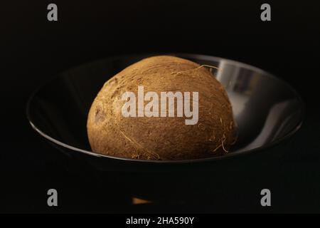 Un cocco organico maturo in un piatto nero, primo piano, isolato su nero. Foto Stock