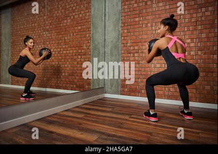 Vista dalla parte posteriore ad una donna africana attraente e sottile che fa esercizi di glute, squatting con la palla medica ginnastica guardando il suo specchio imag Foto Stock