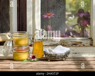 due vasetti di stoccaggio riempiti di unguento di echinacea e una caraffa di vetro di olio vegetale si erigono insieme ad un cesto di metallo e fazzoletti da mano in tessuto su un tavolo di legno di fronte ad una vetrina di officina Foto Stock