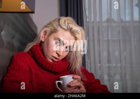 La ragazza caucasica attraente indossa un maglione rosso pesante, è l'inverno ed è al coperto e beve una bevanda calda dalla tazza. Ragazza dell'europa orientale Foto Stock