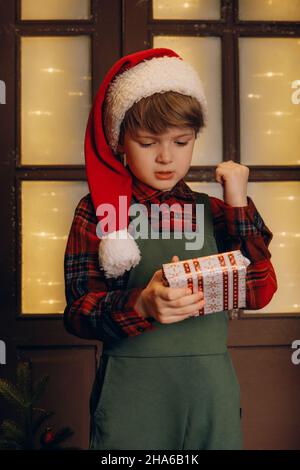 Un ragazzo con i cappelli di Babbo Natale con un cane seduto sul portico di una casa decorata per il natale e scatole con regali nelle sue mani, il concetto di vacanza in famiglia Foto Stock