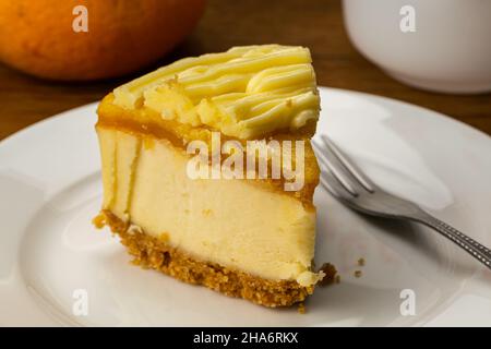 Primo piano di una porzione di deliziosa Cheesecake al limone fatta in casa e forchetta in metallo in ceramica bianca con arancio maturo su sfondo di legno. Foto Stock