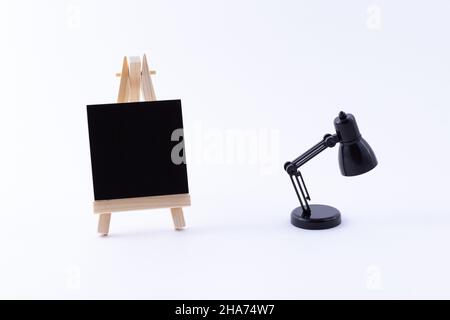 Easel in legno in miniatura con tela quadrata nera vuota per artisti e pittori - Mockup. Mini supporto in legno con tavola da disegno pulita e piccola lampada da tavolo nera su sfondo bianco, Copy Space Foto Stock