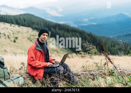 uomo che lavora all'aperto con un computer portatile seduto in montagna. Concetto di lavoro a distanza o stile di vita freelance. Copertura cellulare a banda larga. 5G Internet Foto Stock