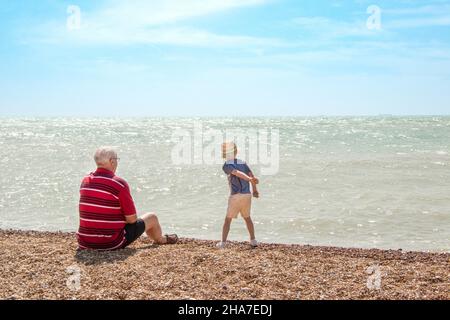 Il ragazzino getta pietre nel mare da una spiaggia di ghiaia nel Kent mentre guardava l'uomo più anziano, probabilmente nonno e nipote in una giornata estiva soleggiata Foto Stock