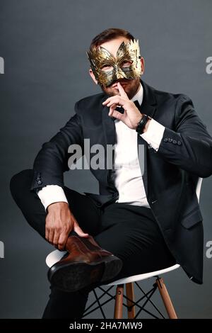 Uomo in tuta da lavoro si siede sulla sedia indossando maschera di carnevale sul suo viso. Ritratto studio su sfondo grigio. Giovane ragazzo di 25-30 anni nasconde il suo volto sotto la maschera. Uomo d'affari astuto e ingannevole. Trickster. Foto Stock