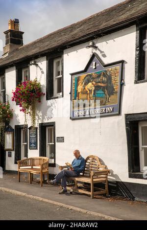 Regno Unito, Cumbria, Allerdale, Keswick, Threlkeld, Uomo che legge al sole sotto il cartello Jennings Brewery Horse e Farrier pub Foto Stock