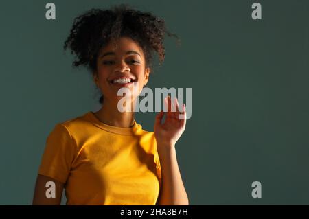 Bella ragazza di etnia afroamericana felice scuro con pelle in maglia gialla che alza la mano nel saluto mentre guarda in macchina fotografica con piacevole sorriso, p Foto Stock