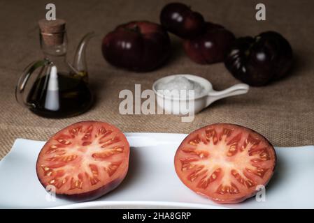 Pomodoro della varietà Mar Azul, diviso a metà e presentato su un piatto, con gli ingredienti per il suo condimento. Foto Stock