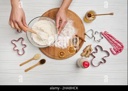 Disposizione piatta. Le donne muovono gli ingredienti secchi con la farina in un recipiente di vetro. Preparare l'impasto di pan di zenzero di Natale. Caramelle, cucchiaio con spezie, bottiglia Foto Stock