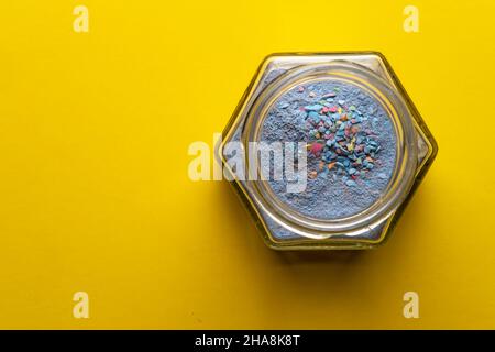 Vaso in vetro aperto con polvere blu e granuli multicolore, su sfondo giallo. Spazio di copia. Vernice colorata allentata. Vista dall'alto. Foto Stock