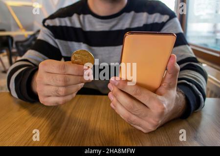 Uomo che tiene smartphone e bitcoin mentre si siede in un moderno caffè. Concetto di investimento di criptovaluta. Acquisto di bitcoin. Foto Stock