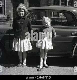 1958, storica, due giovani ragazze, sorelle, si levano in piedi fuori in una strada britannica vicino all'automobile del loro padre, una Renault Dauphine francese, un'automobile posteriore-Engined di economia 4-porta, un successore alla Renault 4CV. Tra il 1957 e il 1961, i Dauphines furono assemblati presso lo stabilimento Renault di Acton a Londra Ovest e uno fu addirittura guidato da HM la Regina. Spaziosa e dallo stile inconfondibile, la Dauphine è stata un successo globale per Renault. Foto Stock