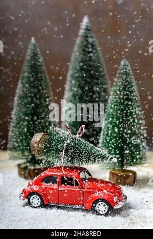 Auto giocattolo rossa con un albero di Natale sul tetto accanto agli abeti mentre sta nevicando su un tavolo di legno. Concetto di celebrare Natale e Capodanno. Foto Stock