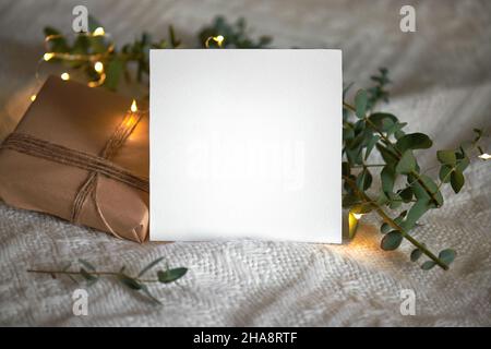 Natale inverno vuoto 5x5 carta mockup quadrato con regalo, eucaliptus foglie e ghirlanda chiara su uno sfondo beige lavorato a maglia, elemento per invito, Th Foto Stock