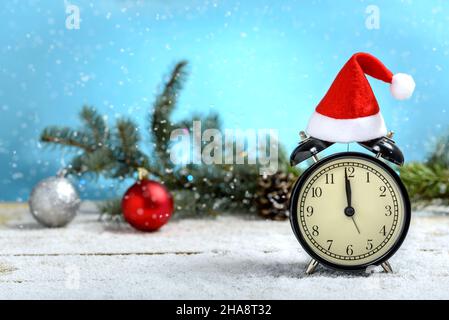Orologio d'allarme vintage con cappello Babbo Natale accanto al ramo di abete mentre sta nevicando su un tavolo di legno. Concetto di celebrare Natale e Capodanno. Foto Stock