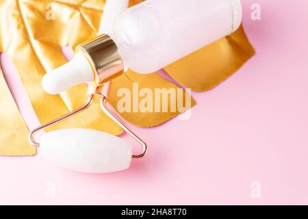 primo piano per la cura della pelle olio cosmetico di rose e rullo facciale in quarzo per il massaggio su foglia di monstera dorata. Procedure di bellezza a casa Foto Stock