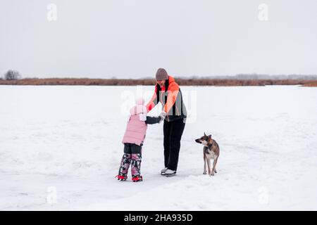 Famiglia a piedi in inverno. La giovane donna insegna alla bambina di pattinare sul lago ghiacciato nella gelida giornata invernale, il grande cane sta giocando nelle vicinanze Foto Stock