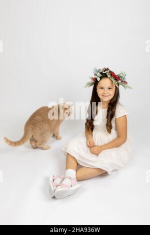 Ragazza sorridente in abito bianco e corona di Natale seduta sul pavimento vicino rosso gatto britannico, isolato su sfondo bianco Foto Stock