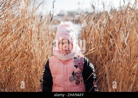 Il bambino è in piedi in erba asciutta. Bambina in rosa abiti invernali si pone sullo sfondo di thicket di canne in giornata di sole in natura Foto Stock