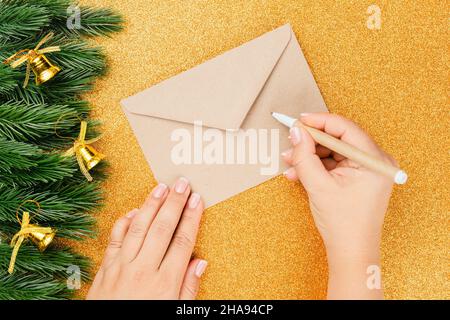 piatto Lay sulle mani donna scrittura lettera in Babbo Natale busta artigianale su sfondo dorato e lucido con decorazioni natalizie Foto Stock