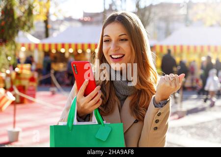 Attraente giovane donna eccitata guardare smartphone e tenere borse shopping a piedi nei mercatini di Natale Foto Stock